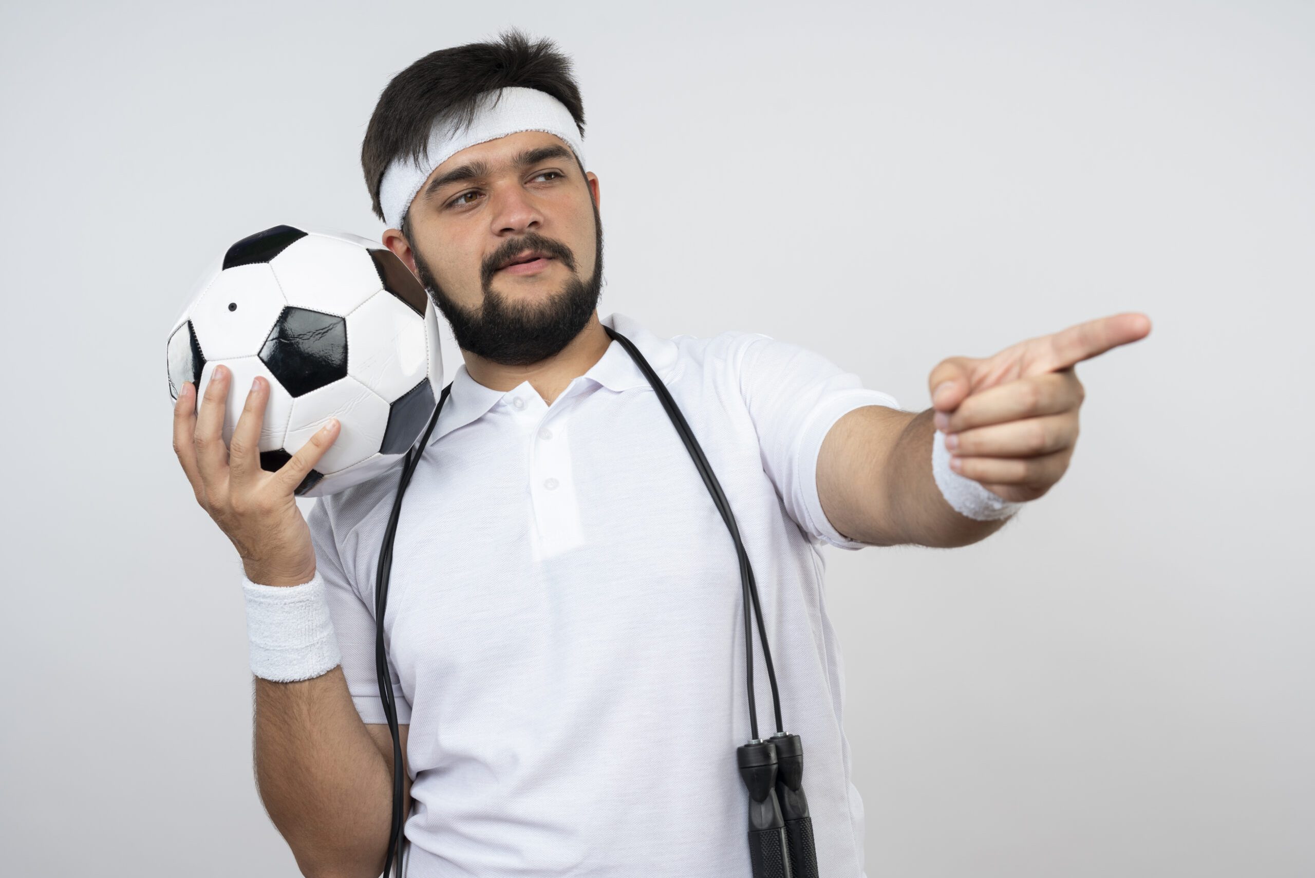 pleased young sporty man wearing headband and wristband with jump rope on shoulder holding ball points at side isolated on white background